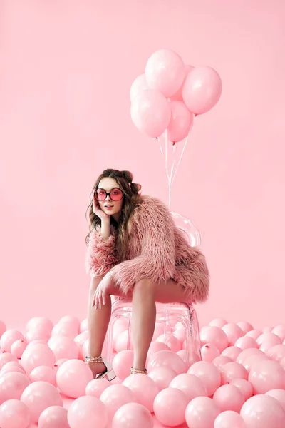 Mujer de moda segura sentada en silla posando sobre muchos globos rosados de fondo —  Fotos de Stock