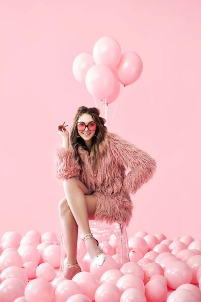 Mulher de moda confiante sentado na cadeira posando em muitos balões rosa fundo — Fotografia de Stock