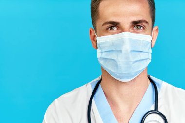 Mavi arka planda tıbbi maske takan yakışıklı erkek doktor portresi.