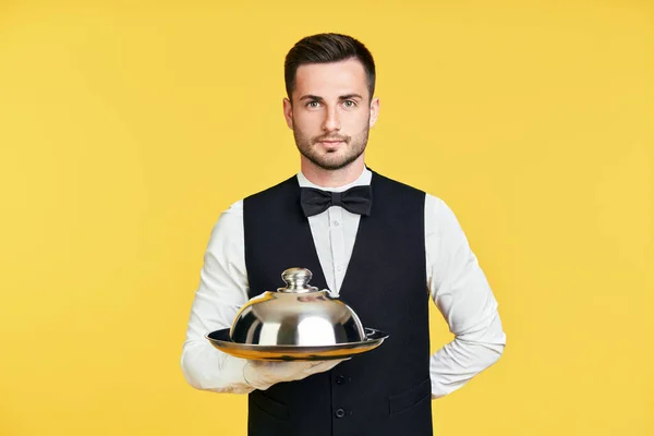 Красивый элегантный официант держит поднос и клош готов служить на желтом фоне — стоковое фото