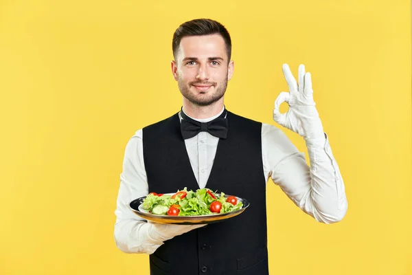 Счастливый улыбчивый официант, держащий тарелку со здоровым салатом, делает хороший знак рукой и пальцами на желтом фоне — стоковое фото