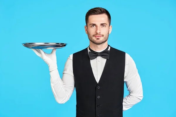 Привлекательный молодой официант держит пустой серебряный поднос на синем фоне — стоковое фото