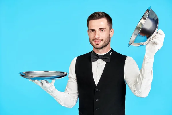 Элегантный молодой официант держит клош над пустым подносом, готовый служить на синем фоне. — стоковое фото
