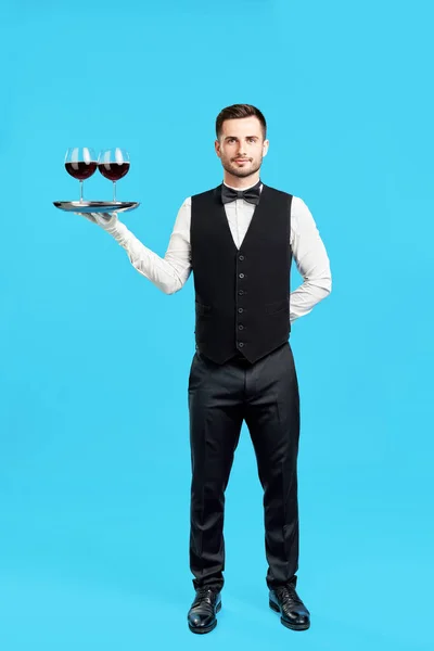 Полнометражный портрет элегантного официанта с бокалами вина на синем фоне — стоковое фото