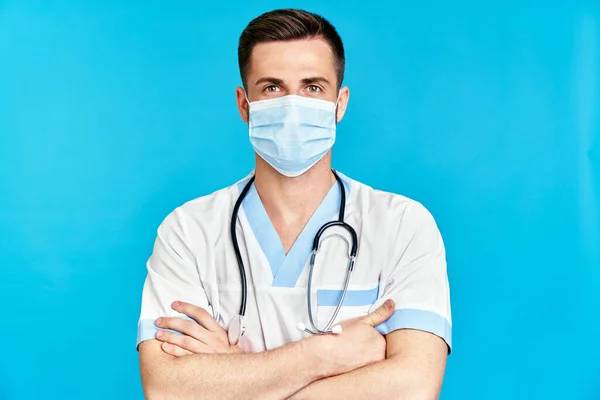 Médico de confianza que lleva máscara médica con los brazos cruzados sobre fondo azul — Foto de Stock
