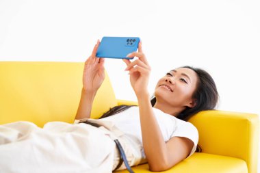 Gülümseyen Asyalı kadın cep telefonuna mesaj yazıyor, sarı koltukta uzanan internet iletişiminden hoşlanıyor.