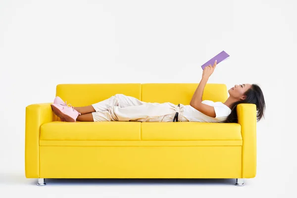 Pretty asian woman reading book lying at cozy yellow couch — Stock Photo, Image