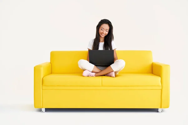 Bella donna sorridente che utilizza il computer portatile seduto sul divano giallo su sfondo bianco — Foto Stock