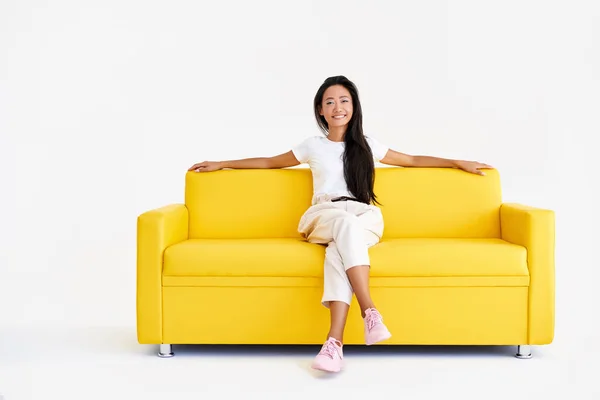 Довольно улыбающаяся азиатская женщина расслабляется на желтом диване на белом фоне — стоковое фото