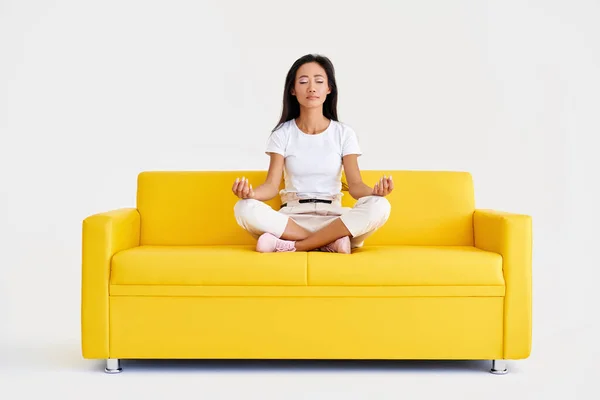 Beautiful young asian woman sitting in yoga lotus pose and meditating on comfortable yellow sofa — Stock Photo, Image