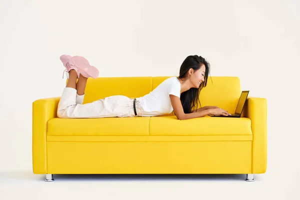 Mujer bastante sonriente usando portátil tumbado en acogedor sofá amarillo sobre fondo blanco — Foto de Stock