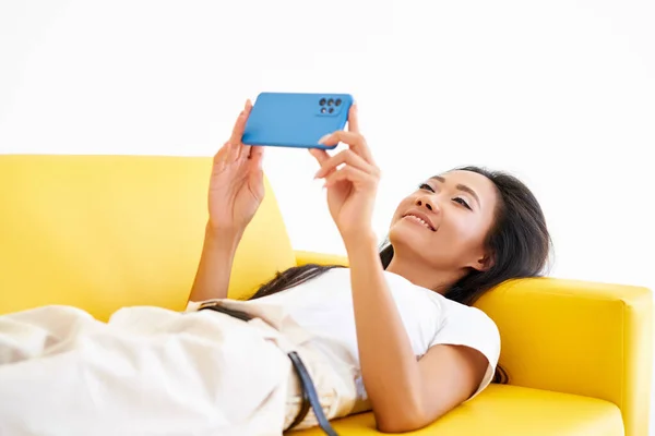 Sonriendo mujer asiática escribe mensaje de texto en el teléfono celular, disfruta de la comunicación en línea acostado en el sofá amarillo — Foto de Stock