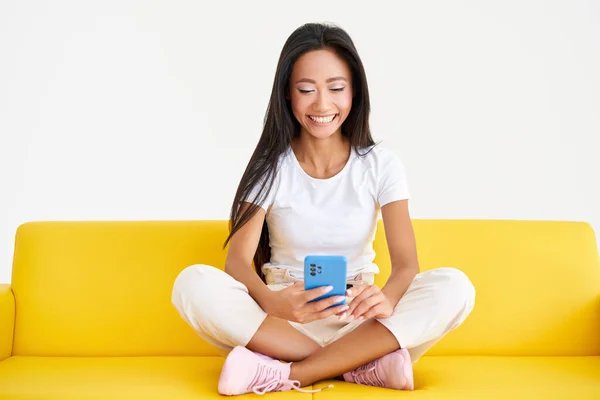 Sonriendo mujer asiática tipos de mensaje de texto, navega por Internet en el teléfono celular sentado en el sofá amarillo — Foto de Stock