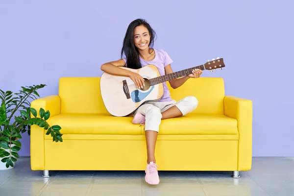 Uśmiechnięta piękna kobieta gra na gitarze siedząc na żółtej kanapie w domu — Zdjęcie stockowe