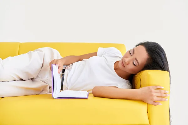 Mooie aziatische vrouw het lezen van boek liggend op gezellige gele bank — Stockfoto