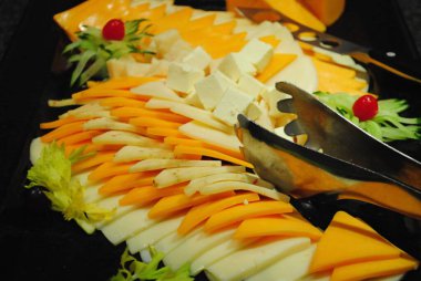 Kahvaltı büfesinde bir çift maşayla farklı peynir dilimleri.