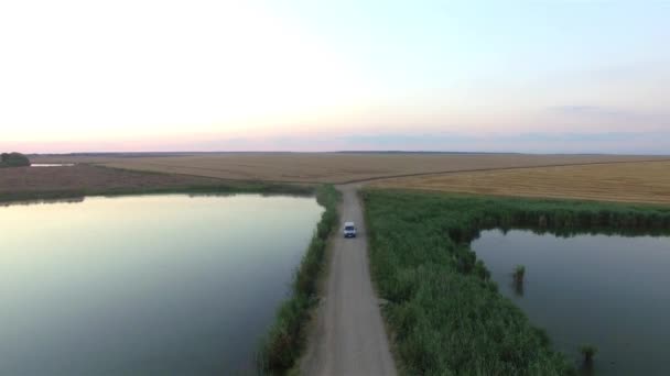 एक निर्जन देश सड़क पर ड्राइविंग करने वाली कार का हवाई दृश्य सूर्यास्त पर दो झीलों से गुजर रहा है — स्टॉक वीडियो