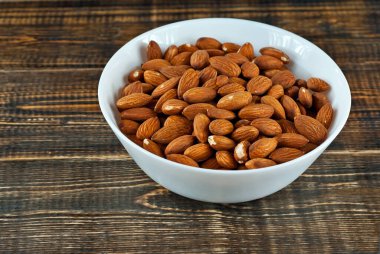 Eski püskü bir tahtanın üzerinde beyaz bir kasede badem. Kahverengi ahşap masada fındıklar.