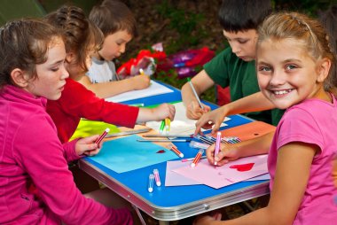 Çocuklar doğada resim yapar. Mavi bir masada bir sürü renkli kağıt ve kalem var. Erkekler ve kızlar dışarıda oynarlar. Parkta resim dersi. Bir çadır kampında dinlen..