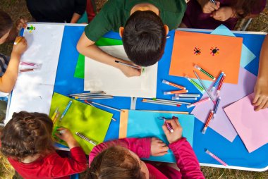 Çocuklar doğada resim yapar. Mavi bir masada bir sürü renkli kağıt ve kalem var. Erkekler ve kızlar dışarıda oynarlar. Parkta resim dersi..