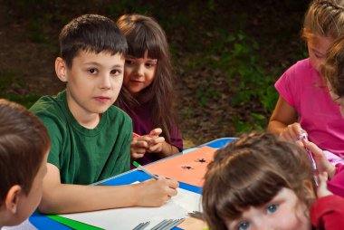 Çocuklar doğada resim yapar. Mavi bir masada bir sürü renkli kağıt ve kalem var. Erkekler ve kızlar dışarıda oynarlar. Parkta resim dersi. Yazın bir çadır kampında dinlen..