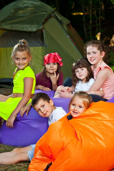 Children rest on a hike. Boys and girls play on an inflatable sofa. Summer vacation in nature. Tents in the background. Games in a tent camp.