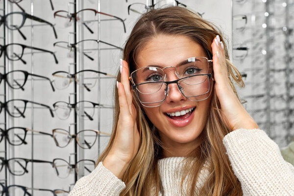 Woman chooses glasses in the store. Blonde in a beige sweater buys glasses. Girl on a background of shop windows with different models of glasses.