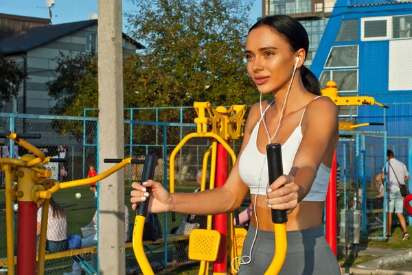 Woman trains on the sports field. Exercise machine for running. Athlete with white headphones and smartphone.