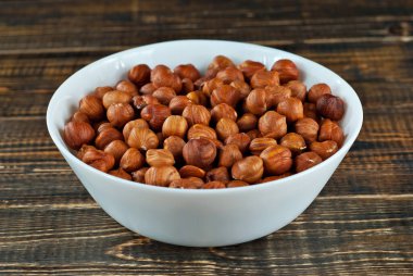 Eski püskü bir tahtanın üzerinde beyaz bir kasede fındık. Kahverengi ahşap masada fındıklar.