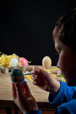 Çocuk bir Paskalya yumurtası boyuyor. Beyaz yumurta elinde, siyah bir arkaplanda..