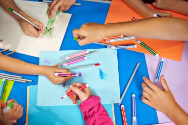 Çocuklar renkli kağıt parçalarına resim çizerler. Eller mavi bir masanın üzerine. Kızlar ve oğlanlar kalemlerle ve keçeli kalemlerle resim yapmayı öğrenirler..