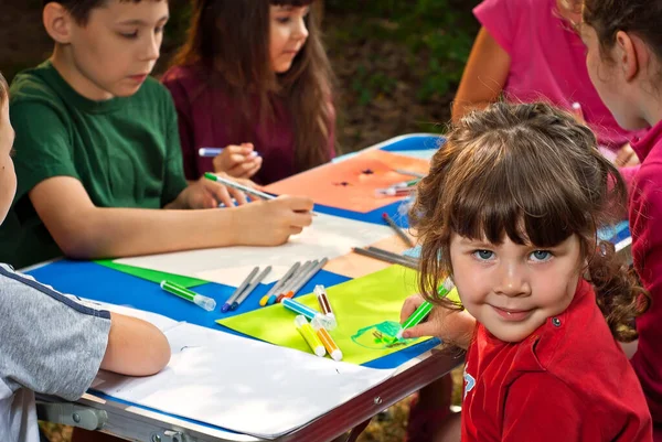 Children draw in nature. Lots of colored papers and pencils on a blue table. Boys and girls play outdoors. Drawing lesson in the park. Rest in a tent camp in the summer.