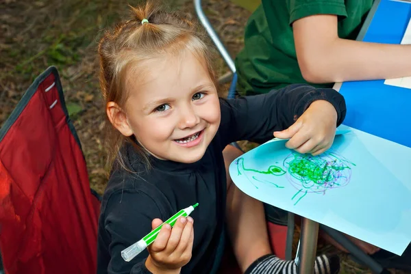 Children draw in nature. Lots of colored papers and pencils on a blue table. Boys and girls play outdoors. Drawing lesson in the park. Rest in a tent camp in the summer.