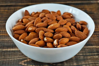 Eski püskü bir tahtanın üzerinde beyaz bir kasede badem. Kahverengi ahşap masada fındıklar.