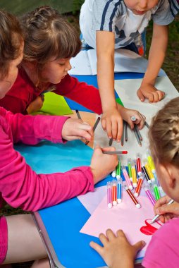 Çocuklar doğada resim yapar. Mavi bir masada bir sürü renkli kağıt ve kalem var. Erkekler ve kızlar dışarıda oynarlar. Parkta resim dersi. Yazın bir çadır kampında dinlen..