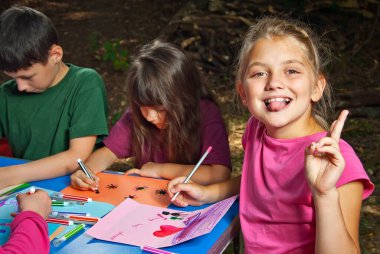 Çocuklar doğada resim yapar. Mavi bir masada bir sürü renkli kağıt ve kalem var. Erkekler ve kızlar dışarıda oynarlar. Parkta resim dersi. Yazın bir çadır kampında dinlen..