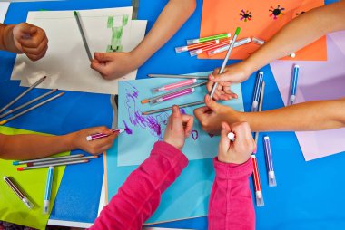 Çocuklar renkli kağıt parçalarına resim çizerler. Eller mavi bir masanın üzerine. Kızlar ve oğlanlar kalemlerle ve keçeli kalemlerle resim yapmayı öğrenirler..