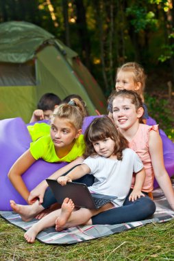 Çocuklar doğada bir dizüstü bilgisayarla oynarlar. Erkekler ve kızlar bir çadır kampında dinleniyorlar. Çocuklar yazın mor bir şişme koltuğa oturur..