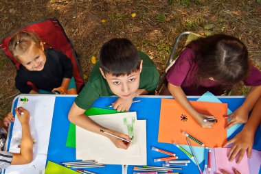 Çocuklar doğada resim yapar. Mavi bir masada bir sürü renkli kağıt ve kalem var. Erkekler ve kızlar dışarıda oynarlar. Parkta resim dersi. Yazın bir çadır kampında dinlen..