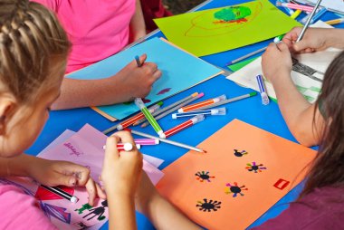 Çocuklar renkli kağıt parçalarına resim çizerler. Eller mavi bir masanın üzerine. Kızlar ve oğlanlar kalemlerle ve keçeli kalemlerle resim yapmayı öğrenirler..