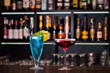 Barda mavi ve kırmızı kokteyller var. Arka planda şişeli alkollü kokteyller. Barın duvarında bulanık alkol şişeleri var..