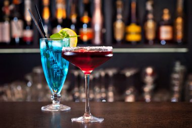 Barda mavi ve kırmızı kokteyller var. Arka planda şişeli alkollü kokteyller. Barın duvarında bulanık alkol şişeleri var..