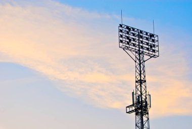 Günbatımı arka planında televizyon antenleri olan bir spot ışığı silueti. Turuncu arka planda stadyum ışıkları için lambalar. Telsiz antenleri projektör kulesine monte edildi..
