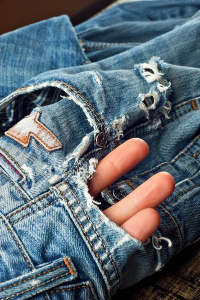 Blue torn jeans on a black background. Hole in the pants close up. The concept of worn clothes and poverty. The hand sticks out of the hole. The man\'s fingers are stuffed into a torn hole.