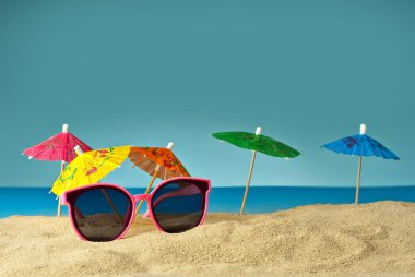 Pembe gözlükler kuma yakın. Kokteyller için arka planda kağıt şemsiyeler olan plajdaki bardaklar. Yaz, deniz ve tatil konsepti.