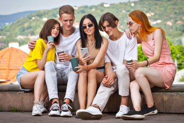 Arkadaşları şehrin arka planında kahve içiyor. Erkekler ve kızlar gülümsüyor. Meydanda güneşli bir günde kaldırımda oturan gençler. Akıllı telefona bakan genç insanlar..
