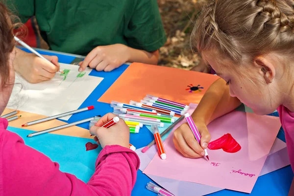 Children draw in nature. Lots of colored papers and pencils on a blue table. Boys and girls play outdoors. Drawing lesson in the park. Rest in a tent camp in the summer.