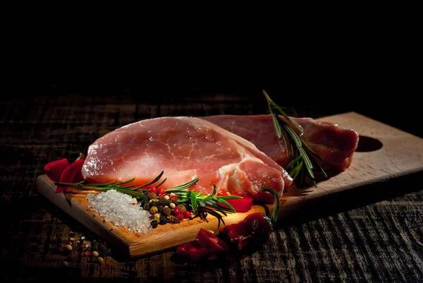 Siyah arka planda tahta bir tahtanın üzerinde çiğ biftek. Eski püskü bir masada kırmızı et. Sanatsal bir efekt olarak dramatik bir ışık. Etin yanında taze biber, tuz ve biberiye..
