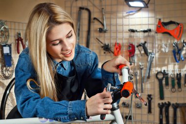 Aletlerin arka planında bisiklet atölyesindeki kadın. Mavi kot gömlekli sarışın bir bisikleti tamir eder. Profesyonel bisiklet servisi kavramı.