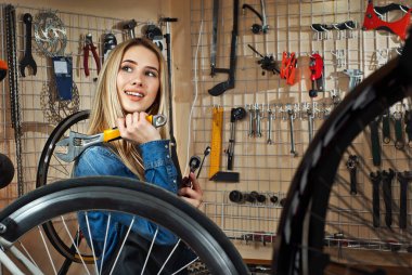 Aletlerin arka planında bisiklet atölyesindeki kadın. Mavi kot gömlekli sarışın bir bisikleti tamir eder. Profesyonel bisiklet servisi kavramı. Elinde ayarlanabilir İngiliz anahtarı olan bir kız..